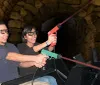 Two people are smiling and wearing 3D glasses seemingly enjoying a ride or attraction where they hold a toy gun together against a backdrop that resembles a stone tunnel