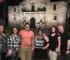 A group of people is posing for a photo at night in front of the historic Alamo mission in San Antonio Texas with one individual holding a Ghost Walk sign suggesting they may be participating in a ghost tour event