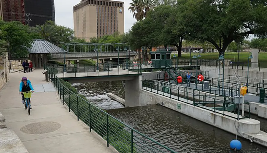 A person is cycling along a riverwalk next to an urban waterway with a bridge and gate mechanism, with pedestrians and greenery in the background.