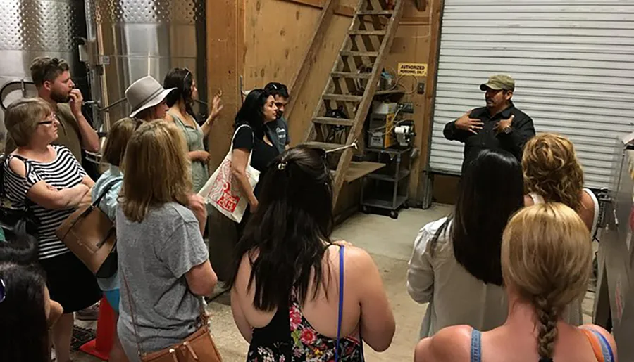 A group of people is attentively listening to a presenter during what appears to be an indoor tour or workshop, possibly in a winery or a brewery, given the stainless steel tanks in the background.