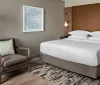 The image shows a neatly arranged modern hotel room with a large bed an armchair a floor lamp and decorative artwork on the wall