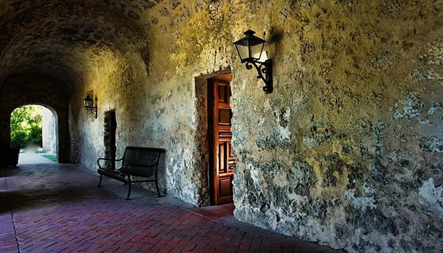The image shows a serene, historic corridor with an arched entrance, a weathered wall, a lantern, a wooden door, and a bench, highlighting the textures and atmosphere of a bygone era.