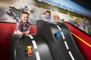 Two children are smiling and playing with toy cars on a curved, oversized race track model with a dynamic scene of racing cars in the background.