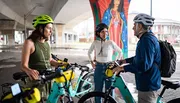 Three cyclists wearing helmets pause for a conversation under an overpass with colorful murals in the background.