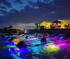 A row of brightly illuminated kayaks casts a vibrant glow on the water at dusk near a waterfront establishment