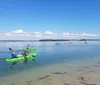 The image shows people in colorful kayaks enjoying a calm day on a clear tranquil body of water under a blue sky dotted with white clouds