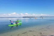 The image shows people in colorful kayaks enjoying a calm day on a clear, tranquil body of water under a blue sky dotted with white clouds.