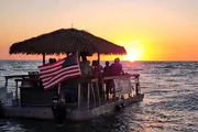 A group of people enjoy a sunset from a thatched-roof boat adorned with an American flag on the open sea.