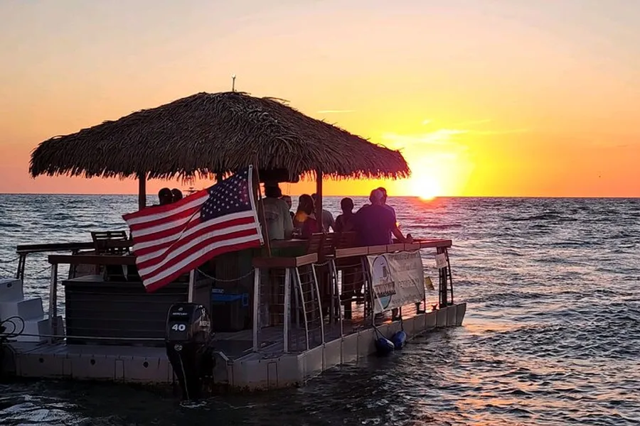 A group of people enjoy a sunset from a thatched-roof boat adorned with an American flag on the open sea.
