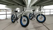 Two fat-tire electric bicycles are parked under a bridge near a body of water.