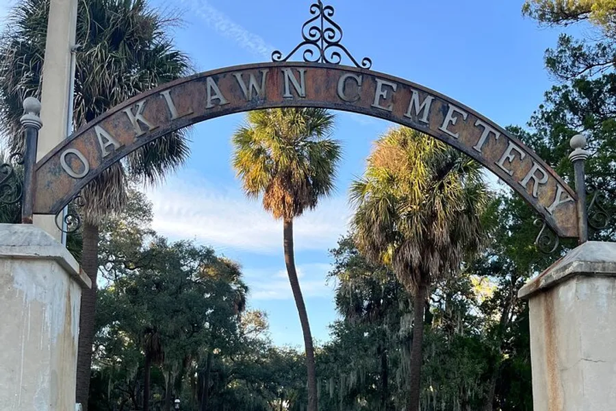 The image shows the rusted iron gate entrance with the sign OAKLAWN CEMETERY, framed by tropical trees under a blue sky.