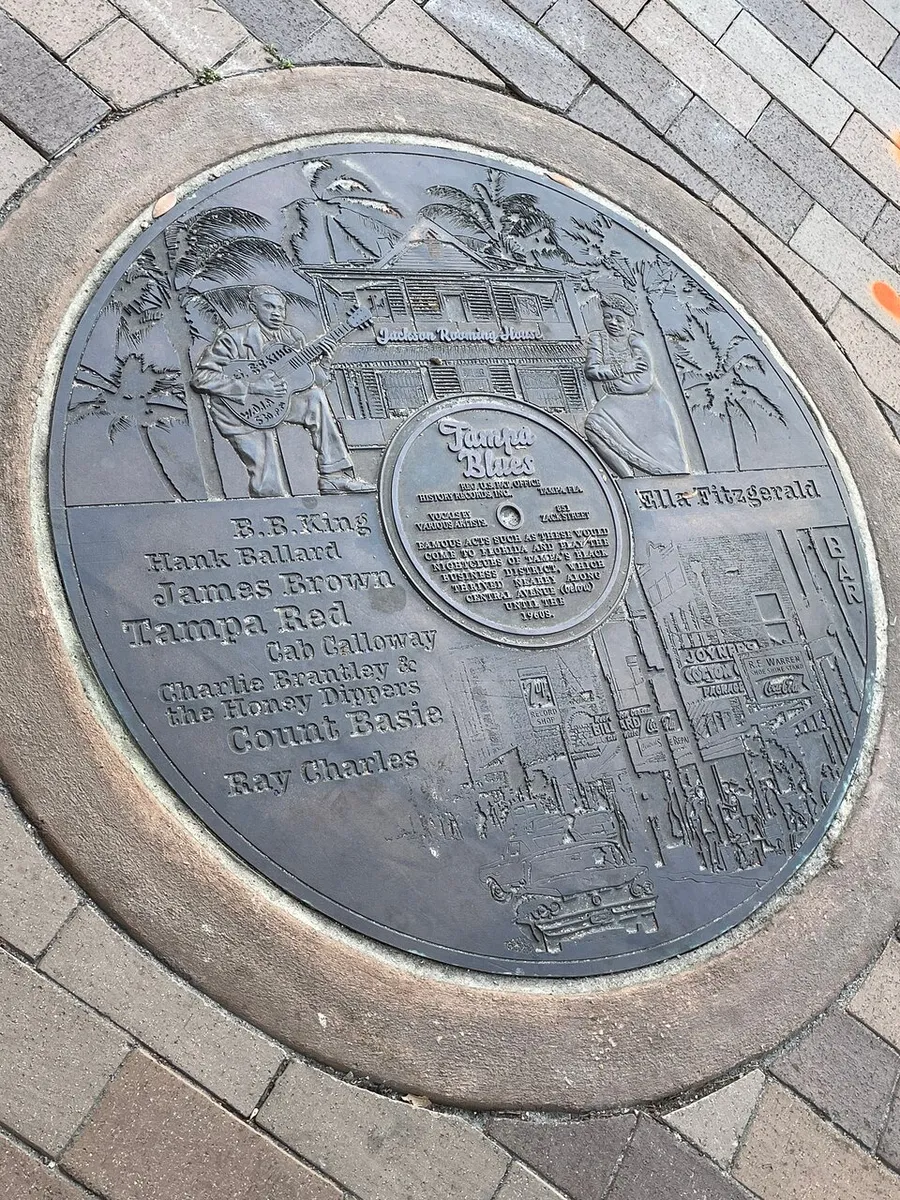 This image features a commemorative plaque embedded in the ground, celebrating artists of the Tampa Blues genre, with intricate engravings and names including B.B. King, James Brown, Ella Fitzgerald, and Ray Charles.