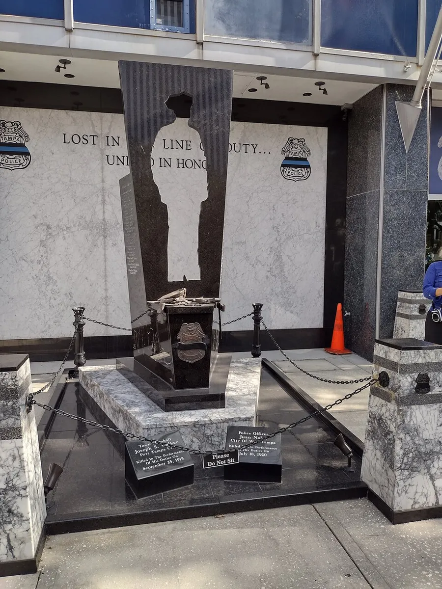 This image shows a solemn memorial for fallen police officers, featuring a black stone monument with an engraved silhouette, inscriptions, and adorned with commemorative plaques and a bronze hat and handcuffs, set against a marbled backdrop with the inscription LOST IN LINE OF DUTY... UNITED IN HONOR.