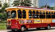 A trolley styled bus adorned with advertisements is parked on a sunny street lined with palm trees.