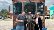 Two people are smiling and posing next to a skeleton figure in front of a trolley-style vehicle that advertises a 