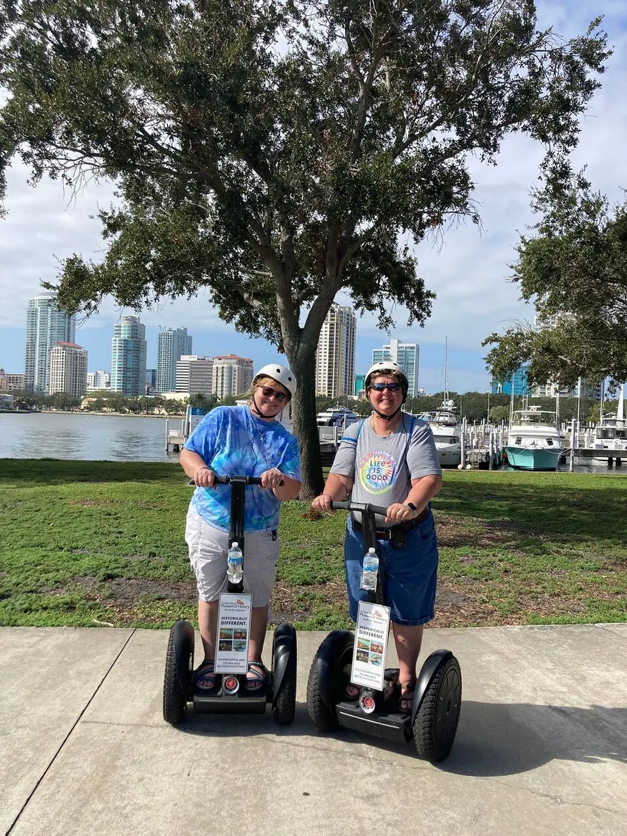 Two people are standing on Segways enjoying a sunny day outdoors with a backdrop of skyscrapers and a marina.