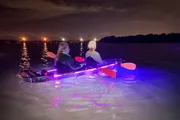 Two individuals are nighttime kayaking with colorful lights illuminating the water around their kayak.