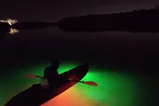 A person is kayaking at night on calm water illuminated by a bright green underwater light.