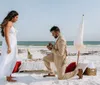 A man is kneeling on one knee on a sandy beach appearing to propose to a woman standing in front of a romantic setup with a table champagne and an ocean backdrop