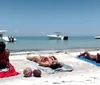 Three people are lying on beach towels sunbathing on a sandy shore with two boats floating nearby in the calm blue water