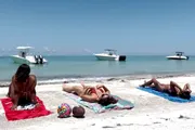 Three people are lying on beach towels sunbathing on a sandy shore, with two boats floating nearby in the calm blue water.