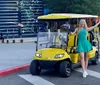 A cheerful person is posing with open arms next to a yellow golf cart on an urban street while other individuals appear in the background