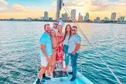 A group of happy friends is posing for a photo on the deck of a sailboat with a scenic view of the water and city skyline at sunset.