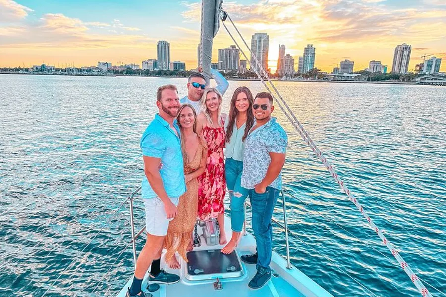 A group of happy friends is posing for a photo on the deck of a sailboat with a scenic view of the water and city skyline at sunset.