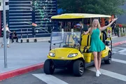 A smiling woman is standing with her arms raised next to a yellow golf cart on an urban street corner.