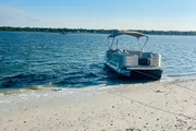 A pontoon boat is moored near a sandy shore with a view of the calm blue water and another boat in the distance.