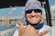 A person wearing sunglasses and a bandana gives a thumbs-up while someone is wakeboarding in the background.