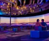 Two individuals are seated on colorful blocks enjoying a vibrant sunset scenery projected on a panoramic screen inside a modern room with artistic decor