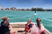 A group of people, including two children wearing life jackets, are on a boat, excitedly watching a dolphin leap out of the water in a sunny coastal setting.