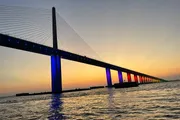 A cable-stayed bridge illuminated with blue lights spans over water against a vibrant sunset sky gradating from yellow to blue.