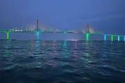 A large, illuminated suspension bridge spans across the water at twilight, its lights reflected on the water's surface.