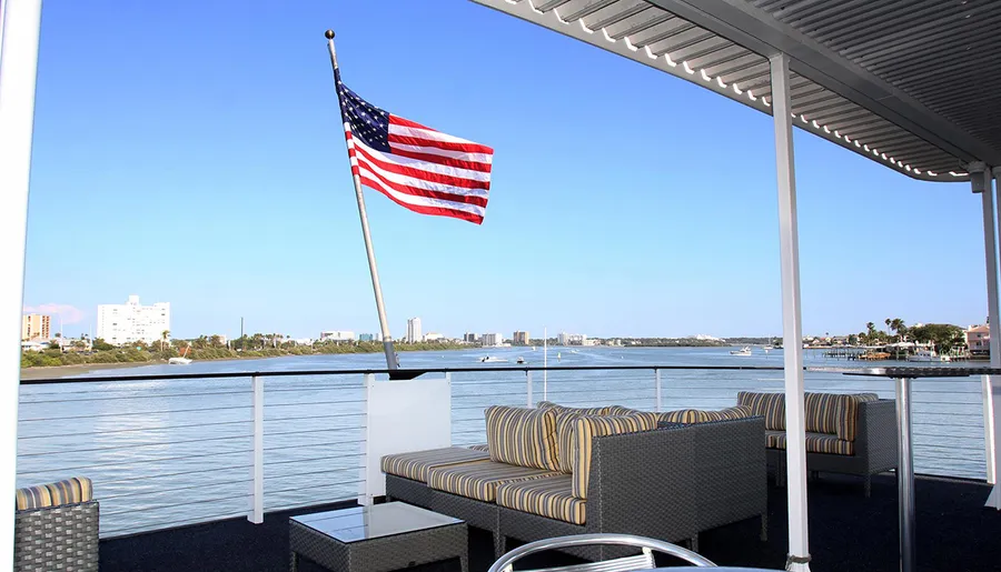 An American flag flies aboard a boat with comfortable seating, overlooking a calm waterfront and a city skyline.