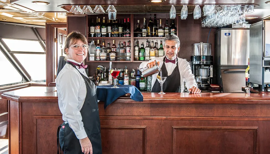 A bartender is pouring a drink beside a smiling colleague at a well-stocked bar.