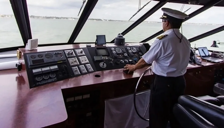 A person in a captain's uniform is standing at the helm of a ship, overseeing a control panel with various navigational instruments.