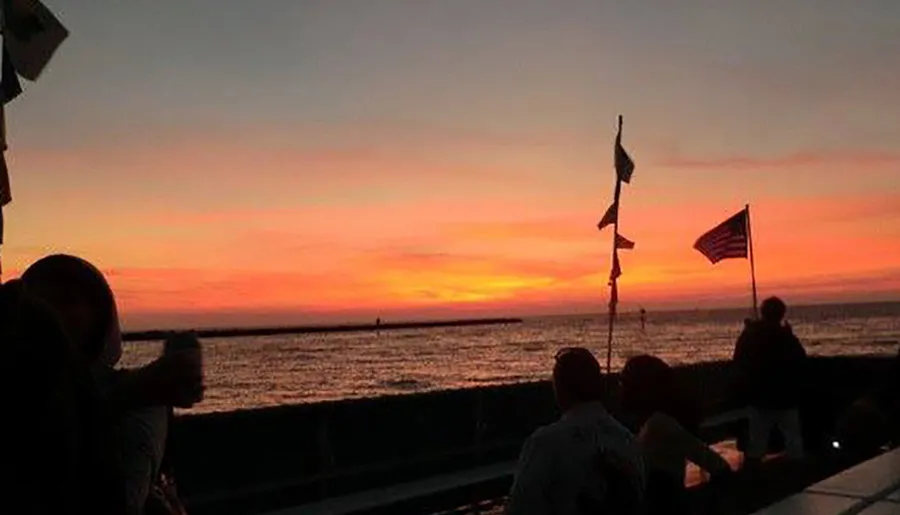 A group of people are enjoying a vibrant sunset by the water, with some flags, including an American flag, silhouetted against the colorful sky.