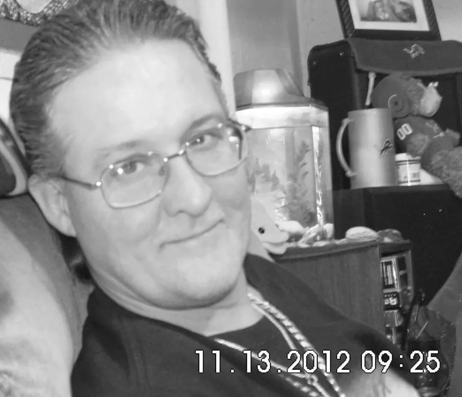 A smiling person with glasses is looking at the camera, with domestic items and plush toys visible in the background, in a black and white photo timestamped 11.13.2012 09:25.