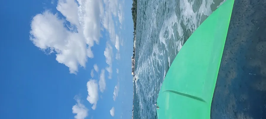 The image shows the edge of a green kayak cutting through blue water with a partly cloudy sky above and a shoreline in the distance.