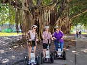 Three people are smiling for the camera while standing on Segways in front of an impressive banyan tree with aerial roots, on a sunny day with a clear blue sky.