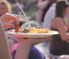 A person is holding a plate of food with a fork and there are other people in the blurry background possibly at an outdoor event
