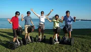A group of five people wearing helmets are smiling and posing on Segways by the seaside.
