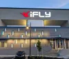 The image shows the modern exterior of an iFLY indoor skydiving facility during dusk or evening with the lit up brand logo prominently displayed above the entrance
