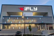 The image shows the modern exterior of an iFLY indoor skydiving facility during dusk or evening with the lit up brand logo prominently displayed above the entrance.