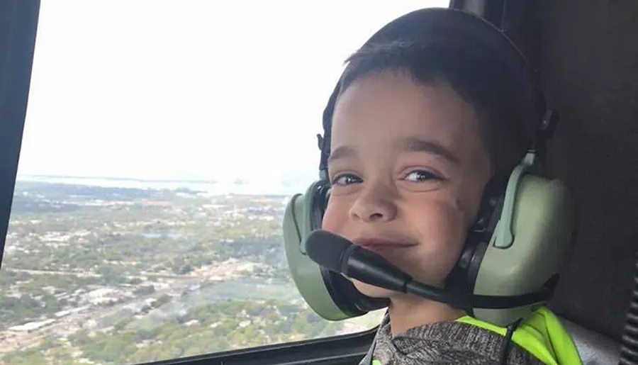 A young child wearing aviation headphones is smiling inside a helicopter with a scenic aerial view in the background.