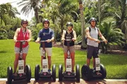 Four individuals are standing outdoors on Segways, wearing helmets and casual summer clothing, with tropical foliage in the background.