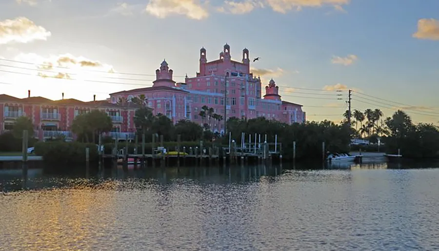 The image shows the pink façade of a grand waterfront building with multiple towers and turrets against a cloudy evening sky, as seen across a body of water with a dock in the foreground.