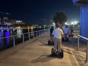 A group of people are riding Segways along a waterfront promenade at dusk with city lights reflecting on the water.